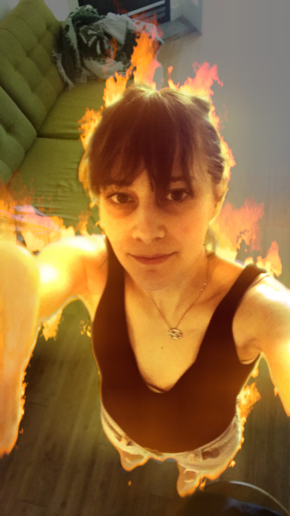 A selfie of Olivia standing in her living room. She is using an Instagram filter that envelops her body in CG flames. She holds the phone camera high above her head, her face smiling softly into her phone. She is a young(ish) white woman and her dark brown hair is pulled back into a ponytail.