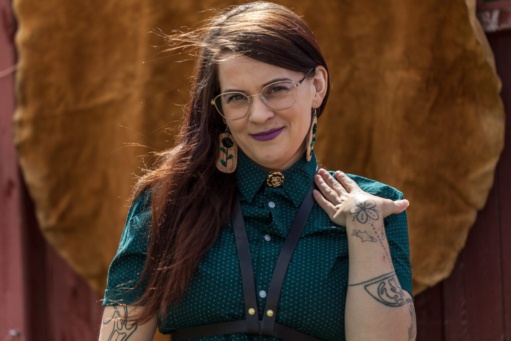 Portrait of Erin Konsmo wearing a teal and white polka-dot blouse, gold-rimmed eyeglasses, floral-design earrings, a gold floral brooch, and black leather suspenders. They are standing in front of a large brown and tan animal hide.