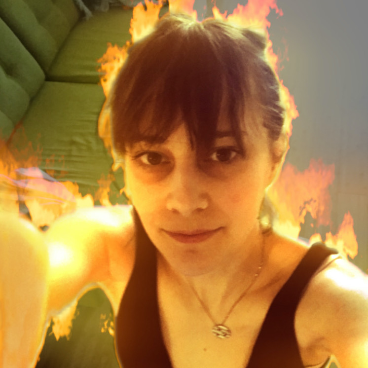 A selfie of Olivia standing in her living room. She is using an Instagram filter that envelops her body in CG flames. She holds the phone camera high above her head, her face smiling softly into her phone. She is a young(ish) white woman and her dark brown hair is pulled back into a ponytail.