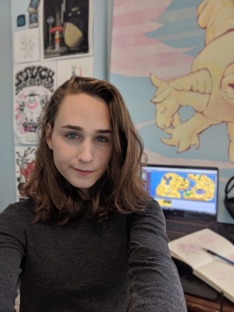 Portrait of Katelyn Hawley wearing a grey sweater sitting in front of a laptop with artwork and posters hanging on the wall behind her.