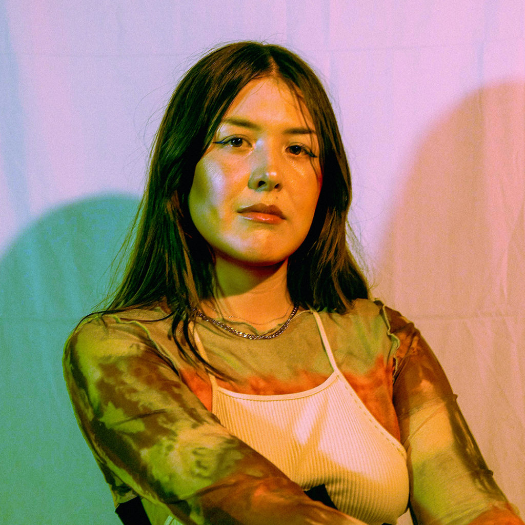 Portrait of Shonee wearing a white tank top over a brown tye-dye shirt with a silver chain necklace. She is sitting in front of a pink and teal backdrop.